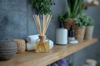 Reed diffuser with bamboo sticks on wooden bathroom shelf with towels, ceramic accessories and green plants creating spa atmosphere at home.