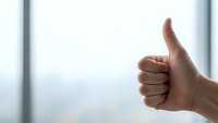 Hand showing thumbs up gesture against bright window background expressing approval, success, and positive feedback for business presentations.
