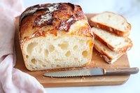 Fresh baked artisan sourdough bread loaf with crispy crust and airy crumb texture, sliced on wooden cutting board with knife, natural light food photography.