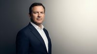Professional middle-aged Caucasian businessman in navy suit and white shirt standing confidently against neutral gradient background for corporate use.