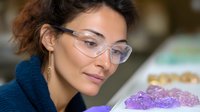Young mixed race woman in protective eyewear examining purple amethyst crystals on white surface, wearing navy sweater, focused expression and natural makeup.