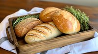 Fresh baked bread rolls with sesame seeds and rosemary sprigs served in rustic wooden tray on white cloth, golden crust artisanal bakery food photography.