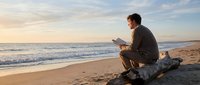 Young man reading book on sandy beach during golden hour sunset with ocean waves and peaceful coastal atmosphere for relaxation and leisure concepts.