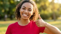 Happy young African American woman with curly hair giving thumbs up gesture outdoors in sunny park setting wearing red shirt with positive expression.