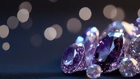 Purple and clear diamonds scattered on dark surface with soft bokeh lights in background creating luxury jewelry atmosphere and sparkling reflections.
