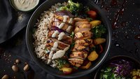 Grilled chicken teriyaki bowl with brown rice, roasted vegetables and creamy sauce served on dark plate. Fresh colorful ingredients create appetizing meal.