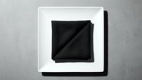 Black fabric napkin or cloth folded on white square plate against gray surface, suitable for restaurant, dining, hospitality, and tableware design projects.