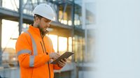 Construction worker in safety gear using digital tablet at building site during golden hour for project management and quality control documentation.