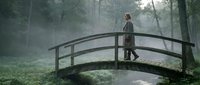 Woman in casual autumn clothing standing on wooden arched bridge in misty forest with stream below, suitable for nature, travel, and lifestyle editorial use.