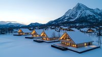 Illuminated modern A-frame chalets nestled in snowy mountain landscape at dusk, cozy winter resort lodges with panoramic windows glowing warmly against white alpine setting.