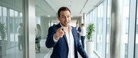 Confident businessman pointing at camera in modern office corridor with glass walls and natural lighting for corporate communication concepts.