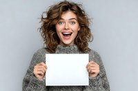 Young woman with curly hair expressing excitement while holding blank white paper against gray background. Perfect for advertising or announcement concepts.
