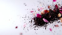 Scattered black tea leaves with pink rose petals and dried flowers creating artistic composition on white background, macro photography with selective focus.