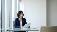 Professional Asian businesswoman in navy suit reviewing documents at modern office desk near windows for corporate presentations and business meetings.