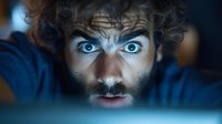 Bearded man with intense blue eyes staring at screen in dark room, illuminated by digital device light, depicting late night browsing or gaming session.