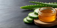 Fresh sliced cucumber with aloe vera leaf and golden honey in glass jar on wooden plate against dark slate background, natural skincare ingredients.