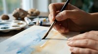 Close-up of hands painting a watercolor artwork with a fine brush on paper, soft blue and golden tones, art supplies in background.