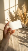 Female hand holding blank white business card mockup in warm sunlight with pampas grass and window shadows creating elegant natural lighting atmosphere.
