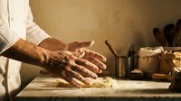 Professional baker kneading fresh dough on wooden table in traditional kitchen. Artisan bread making process with flour dust and warm lighting.