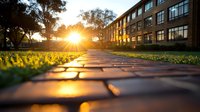 Dramatic sunset light illuminates brick pathway leading to modern office building, with golden sunburst through trees and fresh grass at path edges.