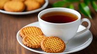 White ceramic cup filled with dark tea served with round butter cookies on saucer, wooden table background with blurred cookies plate and greenery.