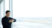 Asian male professional in dark business casual attire checking wristwatch by modern office window with cityscape view. Suitable for corporate, business, time management, and professional lifestyle content.