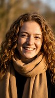 Young Caucasian woman with curly red hair, freckles, and warm smile outdoors in golden autumn light wearing a beige scarf.