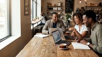 Diverse group of professionals collaborating at wooden table with laptop and documents in modern office cafe setting, discussing project strategy and sharing ideas during business meeting.