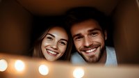 Young diverse couple smiling through cardboard box frame with warm bokeh lights, expressing joy and playfulness during relocation or moving day.