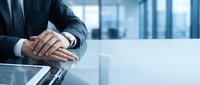 Professional businessman in dark suit with clasped hands on glass conference table in modern corporate office environment for business presentations.