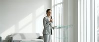 Professional businesswoman in gray suit presenting at glass podium in modern office conference room with natural lighting for corporate meetings.