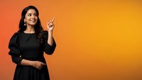 Young Indian businesswoman in black dress pointing upward with finger against orange gradient background for marketing and advertising campaigns.