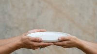 Hands holding empty white plate against neutral beige background, suitable for food styling, product mockups, and culinary presentations.