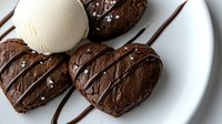 Heart shaped chocolate cookies with vanilla ice cream scoop and chocolate drizzle on white plate, garnished with sea salt flakes for romantic dessert presentation.