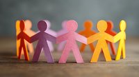 Colorful paper cutout figures holding hands in circle formation on wooden surface against blurred gray background, symbolizing unity and teamwork.