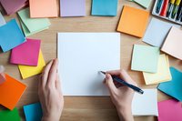 Hands holding pen over blank white paper surrounded by colorful sticky notes on wooden desk for creative brainstorming and planning sessions.