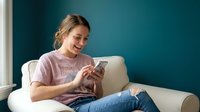 Young Caucasian woman in casual purple t-shirt sitting on white sofa using smartphone, smiling while texting or browsing social media at home.
