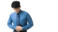 Asian businessman in blue shirt and tie holding stomach with pained expression against white background, depicting digestive discomfort or illness.