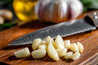 Fresh garlic cloves chopped on wooden cutting board with knife in rustic kitchen setting for cooking and culinary preparation.