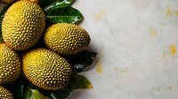 Fresh ripe jackfruit with green leaves on light marble background, top view. Exotic tropical fruit composition with copy space for text.