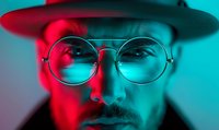 Young bearded man in round glasses and hat illuminated by dramatic red and turquoise neon lighting creating modern cyberpunk atmosphere. Close up portrait with moody lighting.