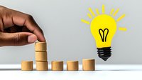 African American hand stacking wooden blocks leading to bright yellow light bulb, representing growth, innovation and business success concept on gray background.