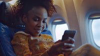 Young African American woman with curly hair smiling while using smartphone during air travel, wearing yellow patterned blouse, perfect for travel, lifestyle, and mobile technology advertising.