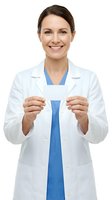 Young female healthcare professional with brown hair and fair complexion wearing white lab coat over blue scrubs, holding blank identification badge, smiling at camera against white background for medical, hospital, and professional identity purposes.