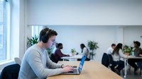 Young professional man with headphones working on laptop in modern open office space with colleagues in background for remote work and productivity concepts.