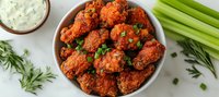Spicy buffalo chicken wings garnished with fresh herbs, served in white bowl with celery sticks and ranch dipping sauce on marble counter. Fresh appetizer for parties.