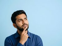 Thoughtful young man with dark hair and beard, South Asian ethnicity, wearing navy blue shirt, hand on chin in contemplative pose against light blue background.