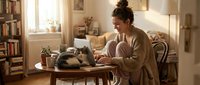 Young Caucasian woman smiling while working on laptop at home with gray cat resting on table, cozy living room interior.