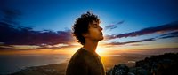 Young man with curly hair looking up at dramatic sunset sky over ocean horizon. Contemplative male silhouette against colorful twilight clouds for inspiration concepts.