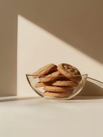 Fresh chocolate chip cookies stacked in clear glass bowl with dramatic diagonal sunlight and shadows on beige background, minimalist food photography style.